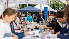 Das Foto zeigt Personen, die links und rechts an Tischen sitzen. Vor ihnen liegen Papier, Stifte und Bastelmaterial auf den Tischen. 