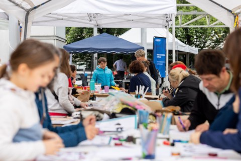 Das Foto zeigt Personen, die links und rechts an Tischen sitzen. Vor ihnen liegen Papier, Stifte und Bastelmaterial auf den Tischen. 
