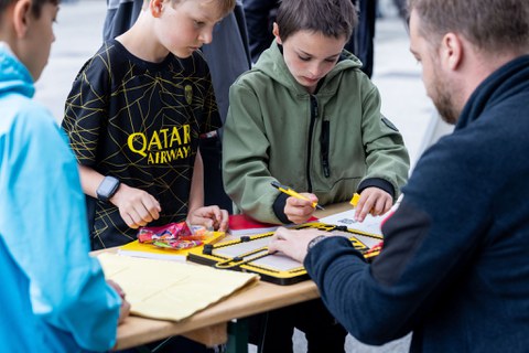 Das Foto zeigt 3 Jungs, die an einem Tisch stehen. An dem Tisch sitzt ein Mann. Einer der Jungs probiert ein Braille-Lineal-Set aus.  