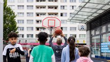 Das Foto zeigt Kinder, die vor einem Basketballkorb stehen, von hinten. Eines der Kinder wirft einen Ball auf den Korb. Im Hintergrund sind Plattenbauten zu sehen. 