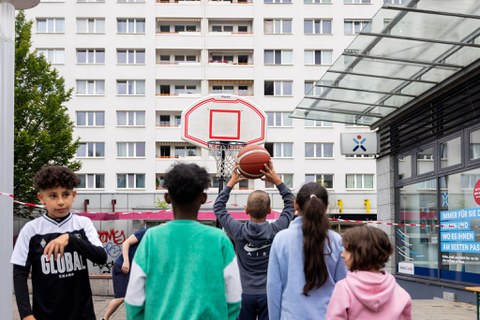 Das Foto zeigt Kinder, die vor einem Basketballkorb stehen, von hinten. Eines der Kinder wirft einen Ball auf den Korb. Im Hintergrund sind Plattenbauten zu sehen. 