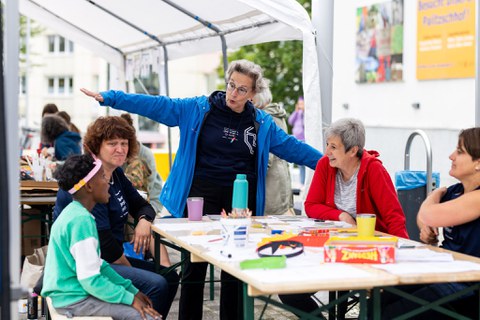 Das Bild zeigt 3 erwachsene Personen und einen Jungen, die an einem Tisch sitzen. Am Tisch steht eine Person mit zur Seite ausgestreckten Armen. 