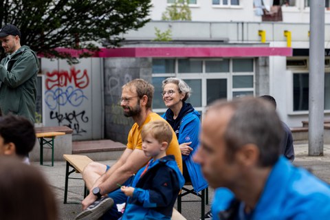 Das Bild zeigt Kinder und Erwachsene, die auf Festzeltbänken sitzen. Im Hintergrund sind mit Graffiti beschmierte Wände zu sehen. 
