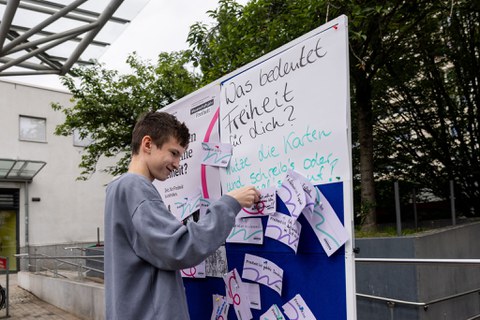 Ein Jugendlicher steht vor einer Pinnwand und steckt eine Karte an der Wand fest. An der Wand steht "Was bedeutet Freiheit für dich?". 