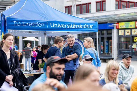 Personen sitzen auf Festzeltbänken und lächeln. Im Hintergrund stehen 3 Personen zusammen und unterhalten sich. 