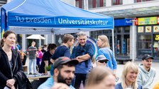 Personen sitzen auf Festzeltbänken und lächeln. Im Hintergrund stehen 3 Personen zusammen und unterhalten sich. 