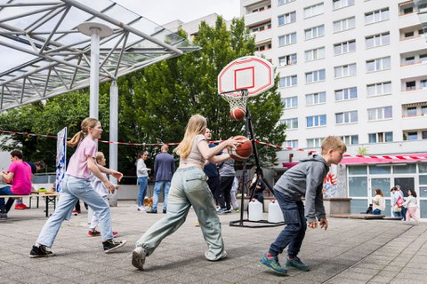 Kinder spielen Basketball. Im Hintergrund sind Plattenbauten zu sehen. 