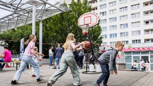 Kinder spielen Basketball. Im Hintergrund sind Plattenbauten zu sehen. 