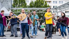 Musiker:innen spielen verschiedene Instrumente. Im Hintergrund sind Plattenbauten zu sehen. 