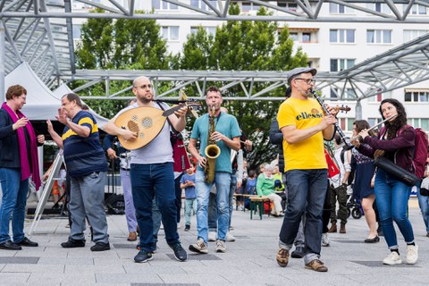 Musiker:innen spielen verschiedene Instrumente. Im Hintergrund sind Plattenbauten zu sehen. 