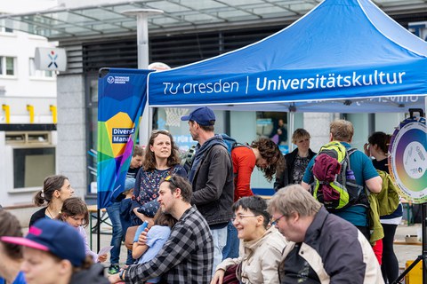 Menschen sitzen und stehen zusammen. Im Hintergrund steht ein Pavillon mit der Aufschrift „TU Dresden | Universitätskultur“.