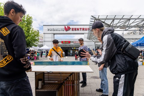 2 erwachsene Männer und 3 Jugendliche spielen Tischtennis mit ungewöhnlichen Schlägern auf einer Platte, die aus verschiedenem Material besteht. 