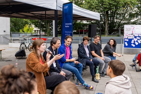 6 Personen sitzen auf Festzeltbänken. Die Person links im Bild hält ein Mikro in der Hand und spricht.