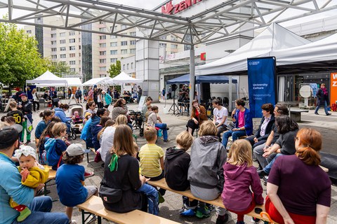 Auf dem Foto sind viele Personen von hinten zu sehen, die auf Festzeltbänken sitzen. 6 Personen sitzen auf Festzeltbänken vorn und den Zuhörern zugewandt.