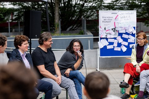 Das Foto zeigt 3 Personen, die auf Festzeltbänken sitzen. Die Person rechts hält ein Mikro und spricht. Im Hintergrund ist eine blaue Pinnwand zu sehen, an der ein Poster mit der Frage "Was wären wir ohne Freiheit?" hängt. 