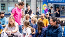 Ein Mann mit pinken T-Shirt steht bei Menschen, die auf Festzeltbänken sitzen. Er hält ein Mikro zu einem Menschen im Publikum. 