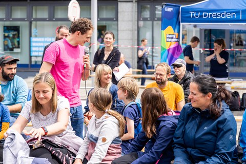 Ein Mann mit pinken T-Shirt steht bei Menschen, die auf Festzeltbänken sitzen. Er hält ein Mikro zu einem Menschen im Publikum. 