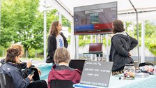 2 Personen stehen neben einem großen Bildschirm und schauen darauf. Im Vordergrund sitzen 3 Personen mit Smartphones in der Hand.