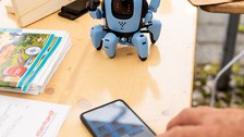 Auf einem Tisch liegt ein Smartphone; eine Hand, die es bedient, ist zu sehen. Auf dem Tisch steht außerdem ein kleiner Roboter. An einem Holzbrett im Hintergrund ist ein Router befestigt. 