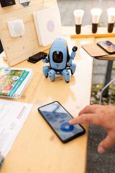 Auf einem Tisch liegt ein Smartphone; eine Hand, die es bedient, ist zu sehen. Auf dem Tisch steht außerdem ein kleiner Roboter. An einem Holzbrett im Hintergrund ist ein Router befestigt. 