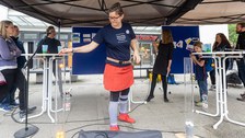 Im Vordergrund steht eine Frau, die einen orangen Ball in eine schmale Glasröhre wirft. Daneben steht ine weitere Röhre, in der 3 weiße Bälle liegen. Links und rechts im Bild stehen jeweils 3 Personen. Zwischen den Gruppen steht eine Frau mit Mikro. 