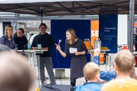 Eine Person mit Mikro schaut und zeigt zu Personen, die auf dem Bild nicht zu sehen sind. Im Hintergrund stehen links 3 Personen an einem Stehtisch neben einer blauen Pinnwand. 