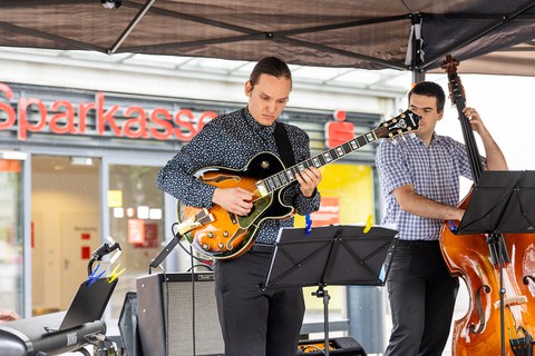 2 Musiker (Gitarre, Kontrabass) spielen unter einem Pavillon. Hinter ihnen sieht man eine Sparkassen-Filiale.