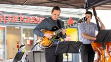 2 Musiker (Gitarre, Kontrabass) spielen unter einem Pavillon. Hinter ihnen sieht man eine Sparkassen-Filiale.