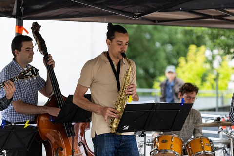3 Musiker (Kontrabass, Saxophon und Schlagzeug) spielen unter einem Pavillon.