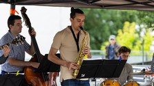 3 Musiker (Kontrabass, Saxophon und Schlagzeug) spielen unter einem Pavillon.