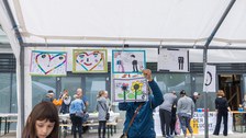 Eine Person hängt gestaltete A4-Blätter an die Querstange eines Pavillons. Links im Bild ist ein Mädchen. Im Hintergrund stehen Personen an verschiedenen Ständen. 