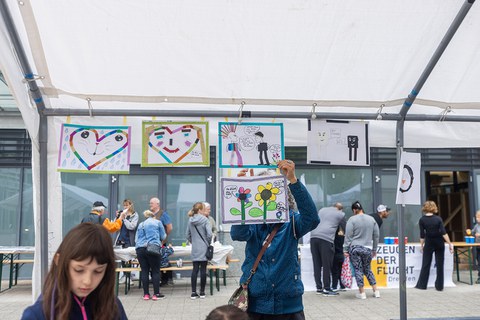 Eine Person hängt gestaltete A4-Blätter an die Querstange eines Pavillons. Links im Bild ist ein Mädchen. Im Hintergrund stehen Personen an verschiedenen Ständen. 