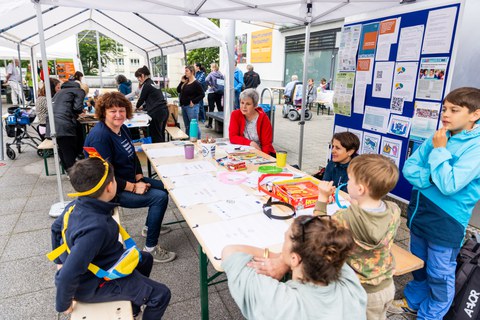 Erwachsene und Kinder sitzen und stehen an einem Tisch unter einem Pavillon. Rechts im Bild steht eine blaue Pinnwand, an der verschiedene Zettel hängen.