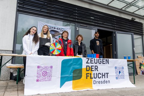 6 Personen stehen hinter einem Tisch, an dem ein Banner mit dem Aufdruck "Zeugen der Flucht2 trägt