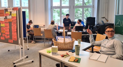 Das Foto teigt einen Seminarraum, in dem an Tischen verteilt Personen sitzen. Im linken Teil des Fotos steht eine Tafel, die mit orangenen, beschriebenen Zetteln behängt ist. 