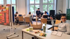 Das Foto teigt einen Seminarraum, in dem an Tischen verteilt Personen sitzen. Im linken Teil des Fotos steht eine Tafel, die mit orangenen, beschriebenen Zetteln behängt ist. 