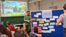 Foto eines Seminarraums. Der rechte Teil des Fotos wird von einer Tafel eingenommen, die mit orangenen, beschriebenen Zetteln behängt ist. Darum stehen Personen, die Zettel aufhängen. In der linke Hälfte des Fotos sitzen Personen um einen Tisch herum. Im Hintergrund ist eine Tafel zu sehen. Darüber wird ein Bild einer Landschaft mit Häusern projiziert. 