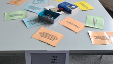 Das Foto zeigt einen quadratischen Tisch von schräg oben. Auf dem Tisch liegen bunte Zettel mit Texten, Stifte und Reißzwecken in einer Dose. An die vordere Kante ist ein Blatt Papier geheftet, auf dem „Philosophieren zu KI“ zu lesen ist. 