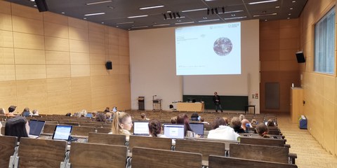 Das Foto zeigt einen Hörsaal. Im Vordergrund befinden sich Menschen mit dem Rücken zum Betrachter, jede:r hat einen Laptop vor sich. Im Hintergrund des Bildes ist eine Person vor einer Power-Point-Präsentation zu sehen, die den sitzenden Menschen zugewandt steht. Die Power-Point zeigt das Bild einer Erdkugel.