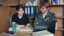 Foto zweier Studierender an einem Tisch. Vor den Studierenden liegen ein Laptop, drei Bücher und Stifte. Beide schauen gemeinsam in ein aufgeschlagenes Buch. 