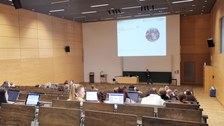 Das Foto zeigt einen Hörsaal. Im Vordergrund befinden sich Menschen mit dem Rücken zum Betrachter, jede:r hat einen Laptop vor sich. Im Hintergrund des Bildes ist eine Person vor einer Power-Point-Präsentation zu sehen, die den sitzenden Menschen zugewandt steht. Die Power-Point zeigt das Bild einer Erdkugel. 