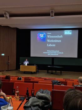 Farbfoto: Vorne befinden sich mehrere Stuhlreihen eines Hörsaals, inkl. einiger Trinkflaschen und Hinterköpfe. Im Hintergrund steht ein Mann am Pult vor einer großen Leinwand. Auf der Leinwand steht: "Wissenschaft, Werkstätten und Labore im Film"