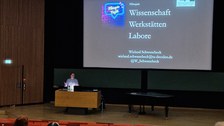 Farbfoto: Vorne befinden sich mehrere Stuhlreihen eines Hörsaals, inkl. einiger Trinkflaschen und Hinterköpfe. Im Hintergrund steht ein Mann am Pult vor einer großen Leinwand. Auf der Leinwand steht: "Wissenschaft, Werkstätten und Labore im Film"