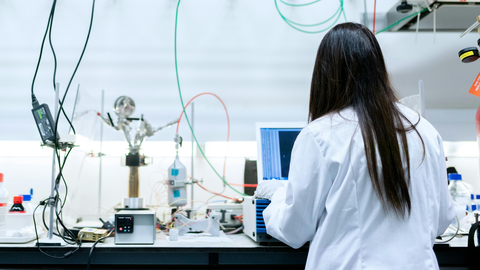 A woman in a white lab coat works on a laptop in a modern laboratory. Scientific equipment, cables, and bottles surround her. Her face is not visible as she is facing away from the camera.
