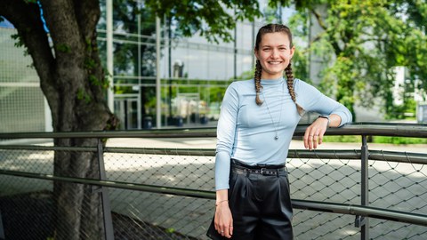 A young woman with braided hair is smiling while leaning against a railing outdoors. She is wearing a light blue long-sleeved top and black shorts. In the background, there is a modern building with glass fronts and trees providing shade.