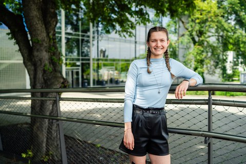 A young woman with braided hair is smiling while leaning against a railing outdoors. She is wearing a light blue long-sleeved top and black shorts. In the background, there is a modern building with glass fronts and trees providing shade.