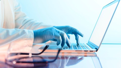 Person in Laborkittel und blauen Schutzhandschuhen arbeitet an einem Laptop; im Vordergrund liegt eine Schutzbrille.