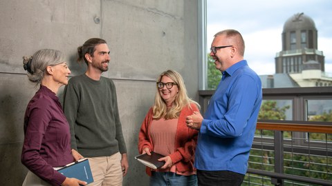 Vier Personen stehen in einem hellen Gebäude vor einer Betonwand und unterhalten sich lächelnd. Zwei halten Notizbücher in der Hand. Im Hintergrund ist durch große Fenster ein markanter Turm sowie etwas Grün zu sehen. Die Atmosphäre wirkt offen, kollegial und freundlich.