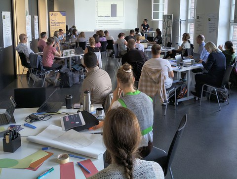Participants of a teaching workshop work at tables with laptops and various materials in a bright seminar room. Paper, pens and facilitation materials are visible in the foreground. In the background, a person presents content related to teaching, projected onto a screen. The group is engaged in discussion and focused work on developing and improving their teaching.
