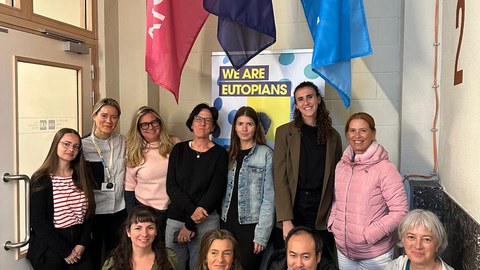 Eine Gruppe von etwa zwölf Personen steht und sitzt für ein Gruppenfoto in einem Innenbereich. Hinter ihnen hängen drei farbige Fahnen und ein Poster mit der Aufschrift „WE ARE EUTOPIANS“. Die Personen lächeln in die Kamera.